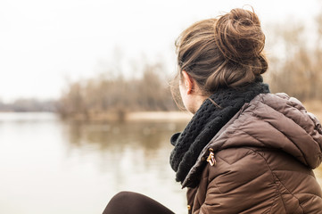 young woman wearing warm coat sitting on the lakeside in early spring
