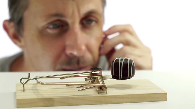 Close up shot of a man looking eagerly at eating a sweet chocolate that is in a mouse or rat trap which indicates that he should not eat it or is on a diet.