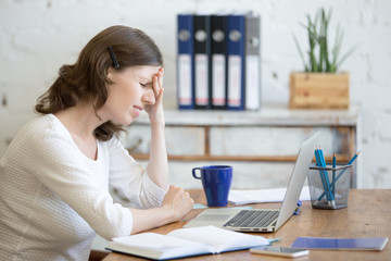 Young stressed businesswoman sitting in front of laptop and touching head with pained expression....