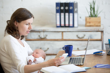 Portrait of young business mom holding her newborn cute babe while working in home office interior, looking at cellphone screen. Serious working mother using mobile phone and nursing new born child