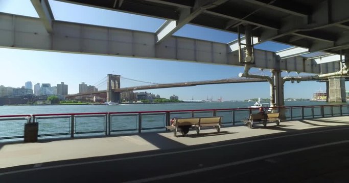 A dolly shot of people walking and jogging on the East River Bikeway under FDR Drive in Manhattan with the Brooklyn Bridge in the background.  	