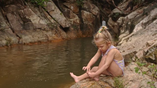 Little Blond Girl Sits Cleans Foot on Stone by Waterfall