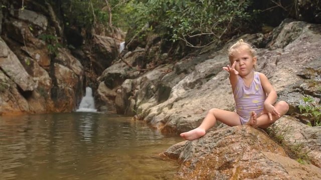 Little Blond Girl Sits Asks for Fruit on Stone by Waterfall