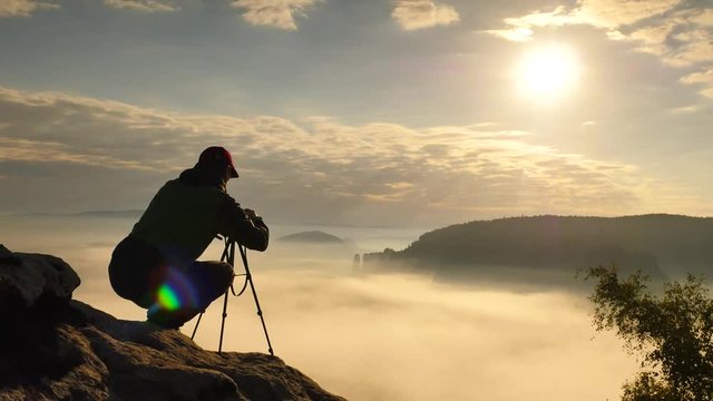 Photographer in green windcheater and black trekking trousers stay at camera on tripod close to cliff edge. Dreamy fogy landscape, blue misty sunrise in a beautiful valley below