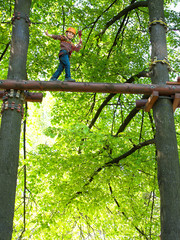 Little boy in helmet and with a safety rope boy goes on a log at a high altitude on the blurred background of trees with yellow and green leaves