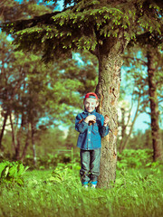 Toned image little boy in a cap and a denim dress standing on the grass and keeps pine cones on blurred background forest