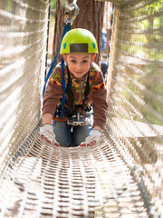 Teenager in helmet and with a safety rope goes on suspension bridge made of rope on the blurred background of trees