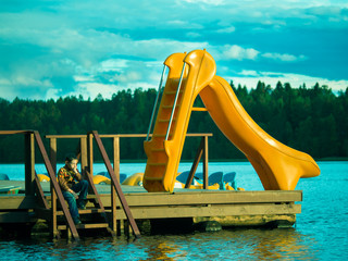 Toned image of a lonely little boy sitting on a wooden pier near the water slide on the background of cloudy sky and forest