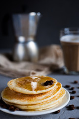 Pancakes decorated with coffee grains on a white plate, moka coffee maker on a vintage tablecloth, a glass of latte on a dark gray background, vertical photo