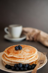 Pancake with blueberry on white plate, golden fork, white cup of coffee on dark gray background, vertical photo