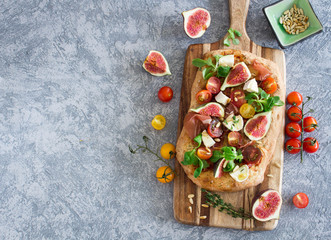 Focaccia with fresh figs, tomatoes, prosciutto and brie cheese on a concrete background, overhead shot