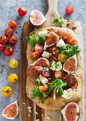 Focaccia with fresh figs, tomatoes, prosciutto and brie cheese on a concrete background, overhead...