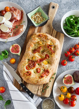 Homemade Italian focaccia with figs, tomatoes, prosciutto, brie cheese, pine nuts and salad on a concrete background surrounded with ingredients