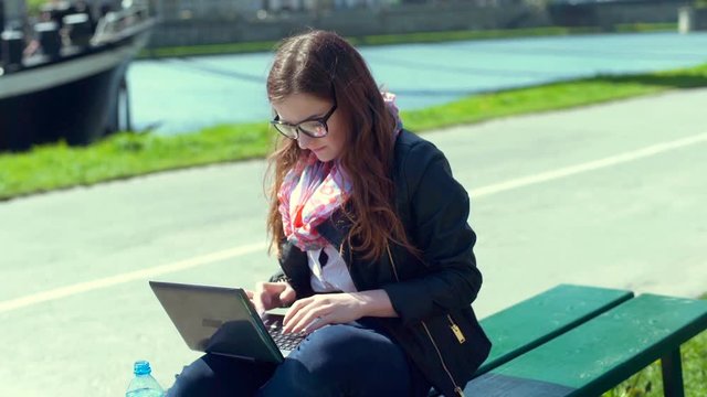 Woman drinking water while using laptop and sitting on the bench
