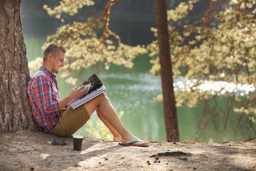 young man student reading a magazine or book on the lake shore