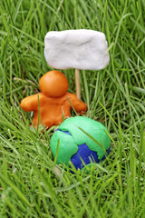 Fototapeta premium Plasticine globe in tall grass. A modelling clay man holds a placard. The concept of Earth Day.