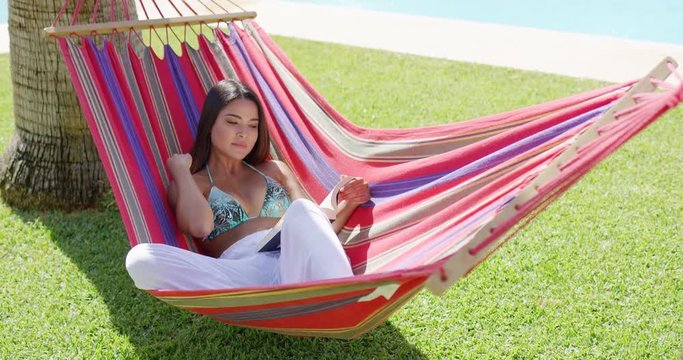 Beautiful single young adult woman in bikini top and white pants seated comfortably in colorful hammock near swimming pool reading a book