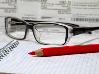 red pencil on a spiral notebook with reading glasses and bills in background