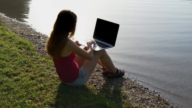 Female Student Sits With Laptop At The Lake Shore