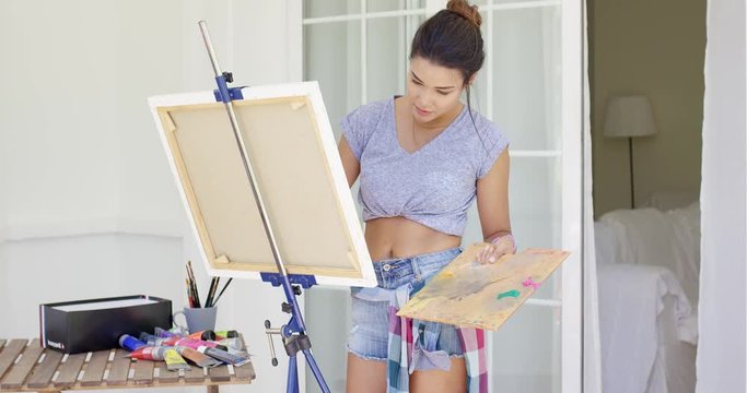 Artistic young woman working on a painting on an outdoor patio looking at the canvas with concentration as she applies the paint