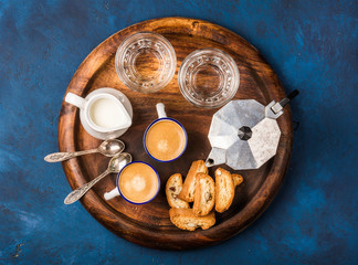 Obraz na płótnie Canvas Coffee espresso in cups with italian cantucci, cookies and milk in jug on wooden serving round board over dark blue painted plywood background, top view, horizontal composition