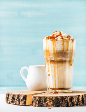 Latte macchiato with whipped cream and caramel sauce in tall glass on wooden board over blue painted wall background, selective focus, copy space, vertical composition