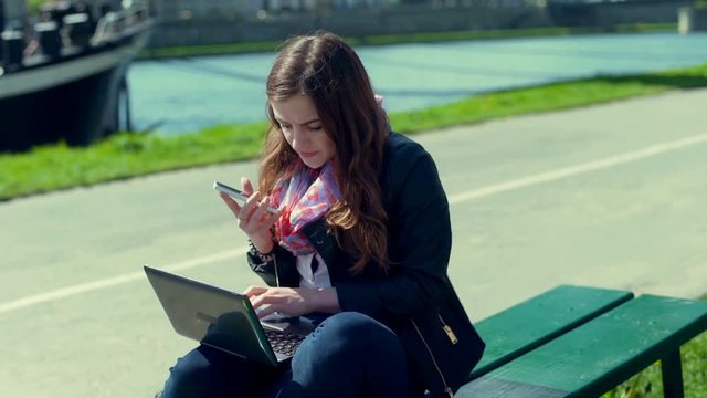 Girl typing on laptop and talking on loudspeaker while sitting on the bench
