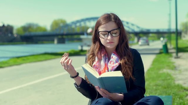 Pretty girl eating a cookie while sitting on the bench and reading book
