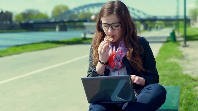Girl eating cookie and typing on laptop while sitting on the bench
