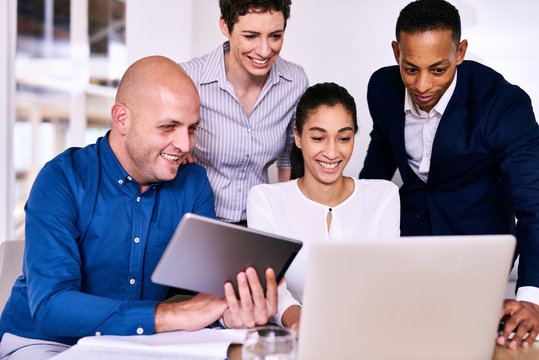 Horizontal image of four business people from diverse backgrounds, of multiethnic complexion unified by working together while looking at a laptop and electronic tablet.