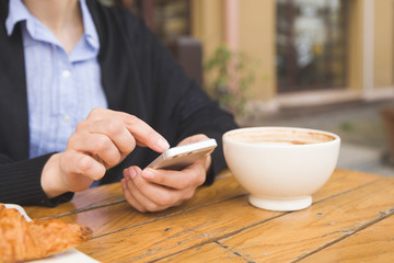 Woman with smartphone sitting in an outdoor cafe and drinking coffee. Breakfast before working day. Cappuccino and croissant on a wooden table. Girl in the cafe.