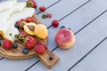 Wooden board with fresh organic fruit and berries: melon, strawberries, blueberries, apricot, peach and red current on grey background. Healthy snack