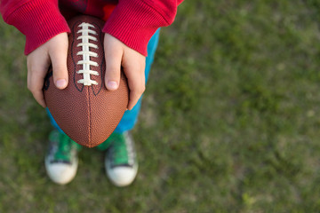Top view on hands of little kid boy holding football on the stadium on a sunny day. Child  ready to throw a football. Sport concept. Sport activities for children outdoors.