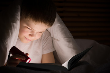 Small kid boy with a smile hiding under a blanket and reading a book with his flashlight in his bedroom at night. Enjoying learning and books.