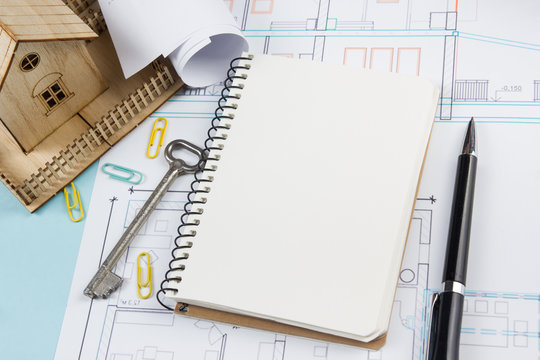 Real Estate Concept. Blank White Notebook On Architectural Desk Table Blueprint Background With Key, Pen, Small House, Office Supplies. Copy Space For Ad Text, Top View.