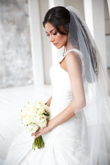 Fototapeta premium Portrait of a beautiful and embarrassment slim bride with the bouquet of peonies in the elegant classic white wedding dress with long veil. Light studio shot
