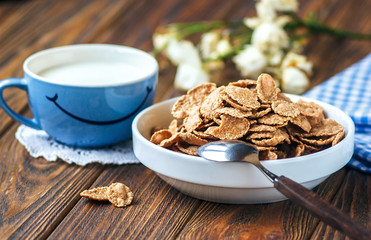 Cereal in white ceramic bowl with spoon on wooden table. Multigrain flakes and cup of milk with smile. Good morning or Have a nice day message concept. Healthy breakfast and diet concept, close up.