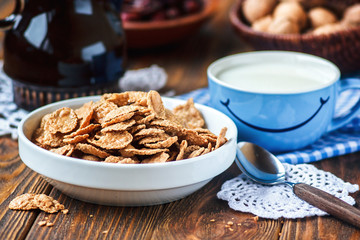 Cereal in white ceramic bowl with spoon on wooden table. Multigrain flakes and cup of milk with smile. Good morning or Have a nice day message concept. Healthy breakfast and diet concept, close up.