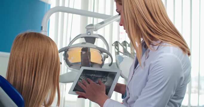 Female dentist showing x-ray image of teeth on digital tablet to her patient and talking to her