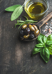 Green and black Mediterranean olives in old cooking spoon with olive oil and herbs over dark rustic wooden background, top view, selective focus, copy space, vertical composition