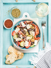 Greek salad with bread slices, oregano, pepper and glass of white wine on blue painted wooden tray over white old wooden background, top view, vertical composition