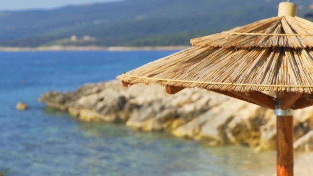 There is a beautiful blue sea seen in the background. A thatched umbrella is in the front of the video on a beach. Close-up shot.
