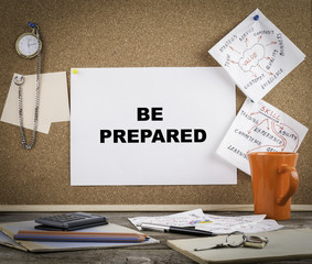 BE PREPARED. Text on cork board. Wooden office desk with a big m
