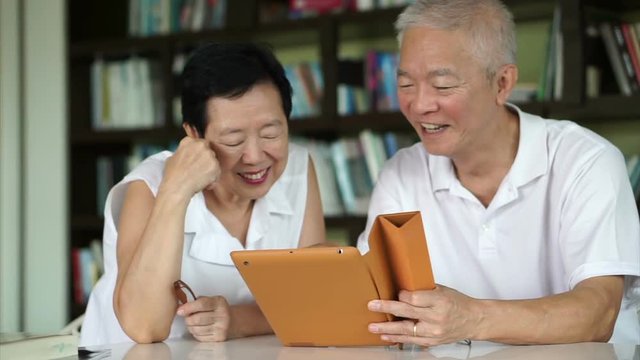 Asian senior couple reading news together from tablet in small library. Learning and study concept