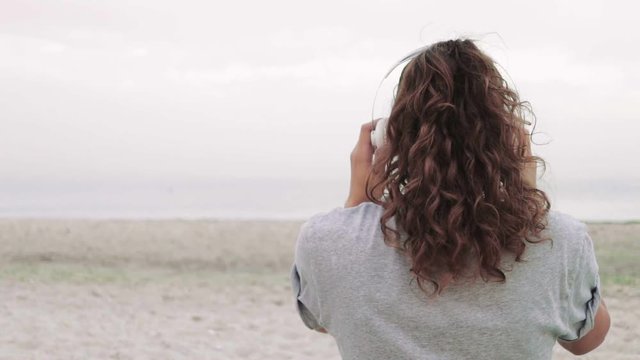 Young curly brunette wears headphones and listens to music on the beach, slow motion, view from the back
