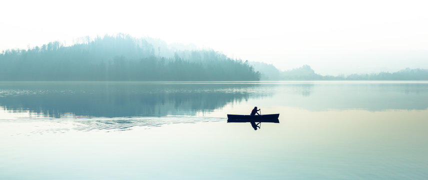 Fog over the lake. Silhouette of mountains in the background. The man floats in a boat with a paddle.