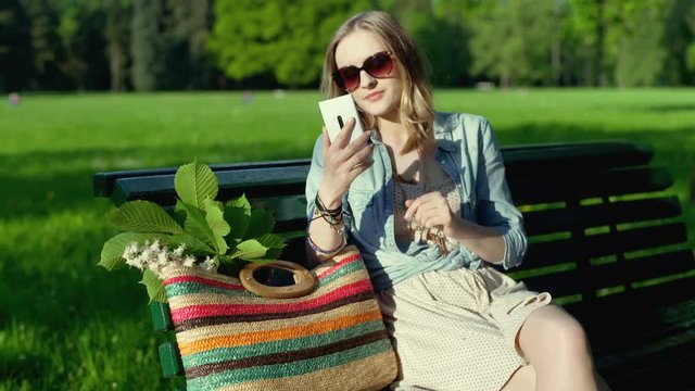 Pretty girl sitting on the bench in the park and texting on smartphone
