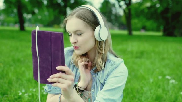 Pretty girl wearing headphones and doing selfies on tablet in the park
