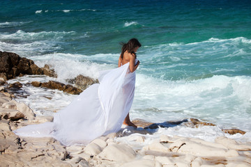 Fototapeta na wymiar Woman in white cape dress stands on the rocky beach on the sea