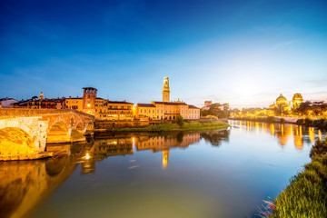 Obraz premium Verona cityscape view on the illuminated riverside with historical buildings and towers on the sunset